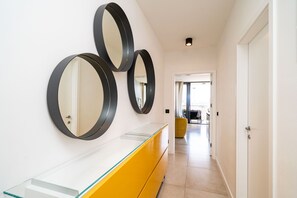Een gang met een glazen en geel ogend wastafelmeubel, drie spiegels aan de muur, en een deur die leidt naar een andere kamer.