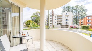 A balcony with a table and chairs, a glass door, and a view of a residential area with buildings and a road. - Seashells, 2,21-23 Magnus Street -Unit with Air con, complex lift and pool, close to town (Nelson Bay)