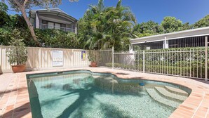 A swimming pool with a curved edge, surrounded by a white fence, and a building with a curved roof in the background. - Seashells, 2,21-23 Magnus Street -Unit with Air con, complex lift and pool, close to town (Nelson Bay)