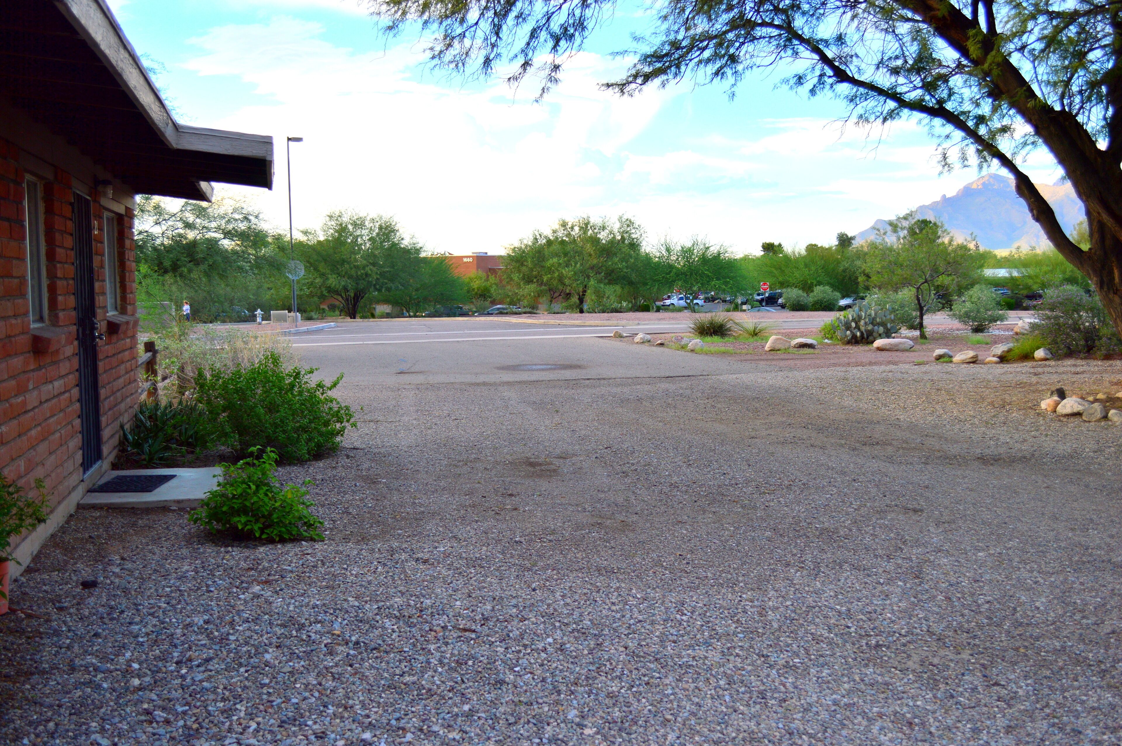 A parking lot with a building on the left, surrounded by gravel and small plants.