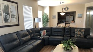 Ein Wohnzimmer mit einem schwarzen Leder-Polstersofa, einem runden Couchtisch und einem Essbereich, im Hintergrund ist eine Küche zu sehen.