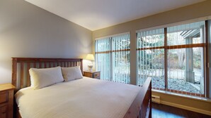 A bedroom with a large bed, wooden headboard, a nightstand with a lamp, and a window with a view of trees and a patio. - Stoney Creek Northstar - Renovated, Ground Floor Condo in Whistler Village. (Whistler)