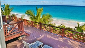 Une terrasse en bois avec des chaises blanches donnant sur une plage aux eaux turquoise et cristallines, bordée de palmiers.
