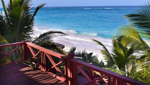 Une terrasse en bois avec des garde-corps rouges, donnant sur une plage où l’eau est d’un bleu limpide et où l’on peut admirer des palmiers.