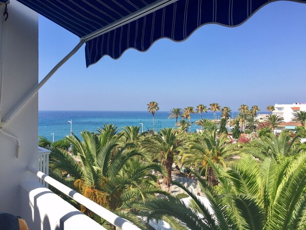 Un balcón con vista al mar, palmeras y un edificio al fondo.