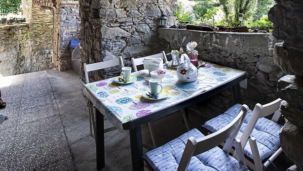 Un arc en pierre mène à une table dressée avec une nappe à motifs floraux, des chaises et une théière.