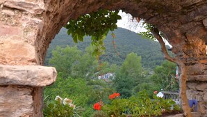 Un arc en pierre encadre une vue sur un jardin luxuriant, où poussent différentes plantes et fleurs, et mène à une colline où se trouve un bâtiment au loin.