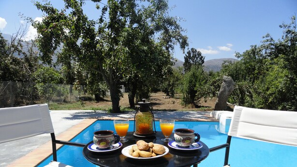 A poolside table with breakfast items, two white chairs, and a view of trees and mountains. - Zeus House Gaia (Lasithi Plateau)