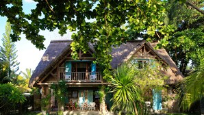 Une maison à deux étages, dotée d’un toit de chaume, entourée d’une végétation luxuriante et d’une pelouse bien entretenue.