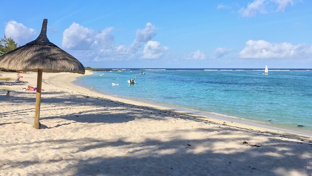 Une plage avec un parasol en chaume, une eau d’un bleu limpide et quelques bateaux à l’horizon.