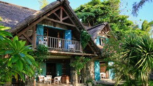 Une maison en bois à l’allure rustique, dotée d’un balcon et entourée d’une végétation luxuriante et de plantes tropicales.