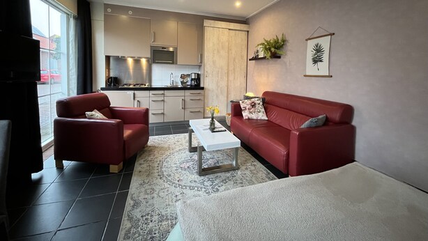 A modern living room with red sofas, a coffee table, and a kitchen in the background. - VRIJ van 29 augustus tot 5 september .2 pers studio 5 min lopen  het strand (Westkapelle)