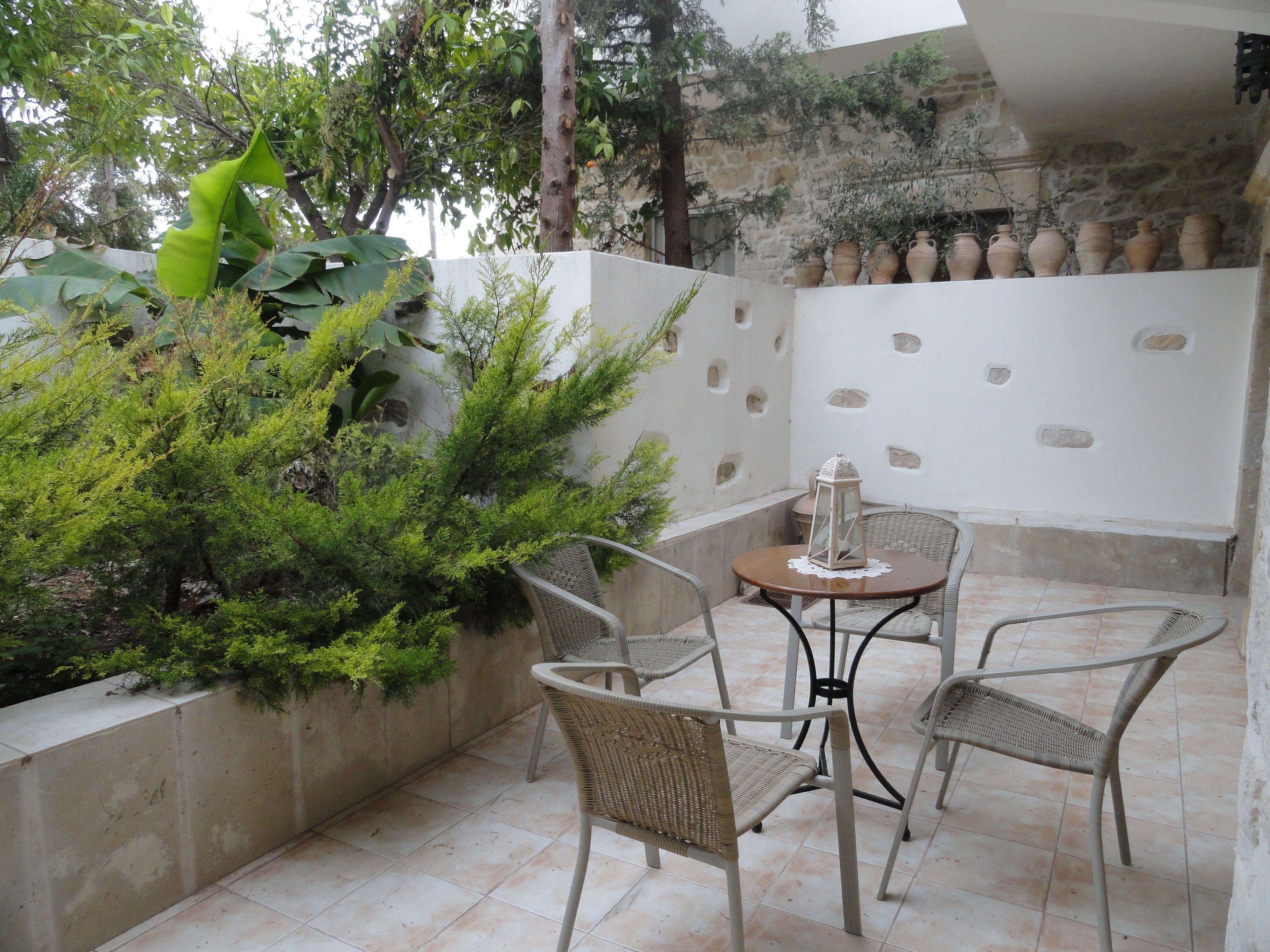 A patio area with wicker chairs and a table, surrounded by greenery and stone walls.