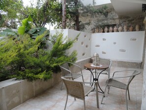 A patio area with wicker chairs and a table, surrounded by greenery and stone walls. - Villa Arokaria (Faistos)
