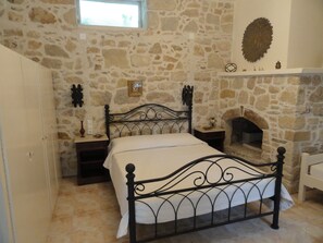 A bedroom with a stone wall, a fireplace, a bed with a metal frame, and a small bedside table with a lamp. - Villa Arokaria (Faistos)
