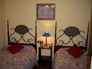 A room with two beds, each with a headboard and a red pillow. There is a small table with a lamp between the beds.