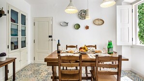 Ein Essbereich mit einem Holztisch, gedeckt für vier Personen, umgeben von Stühlen. Es gibt Pendelleuchten, eine weiße Tür und ein Fenster mit Grünpflanzen draußen.