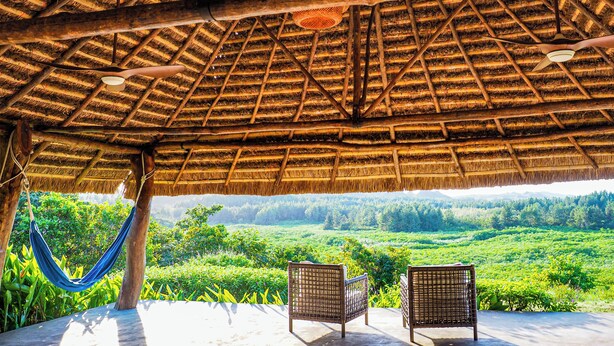 Estructura con techo de paja, muebles de mimbre e iluminación colgante, con vistas a un paisaje verde y exuberante.