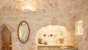 A stone-walled room with a bed, a mirror, a sink, and a window.