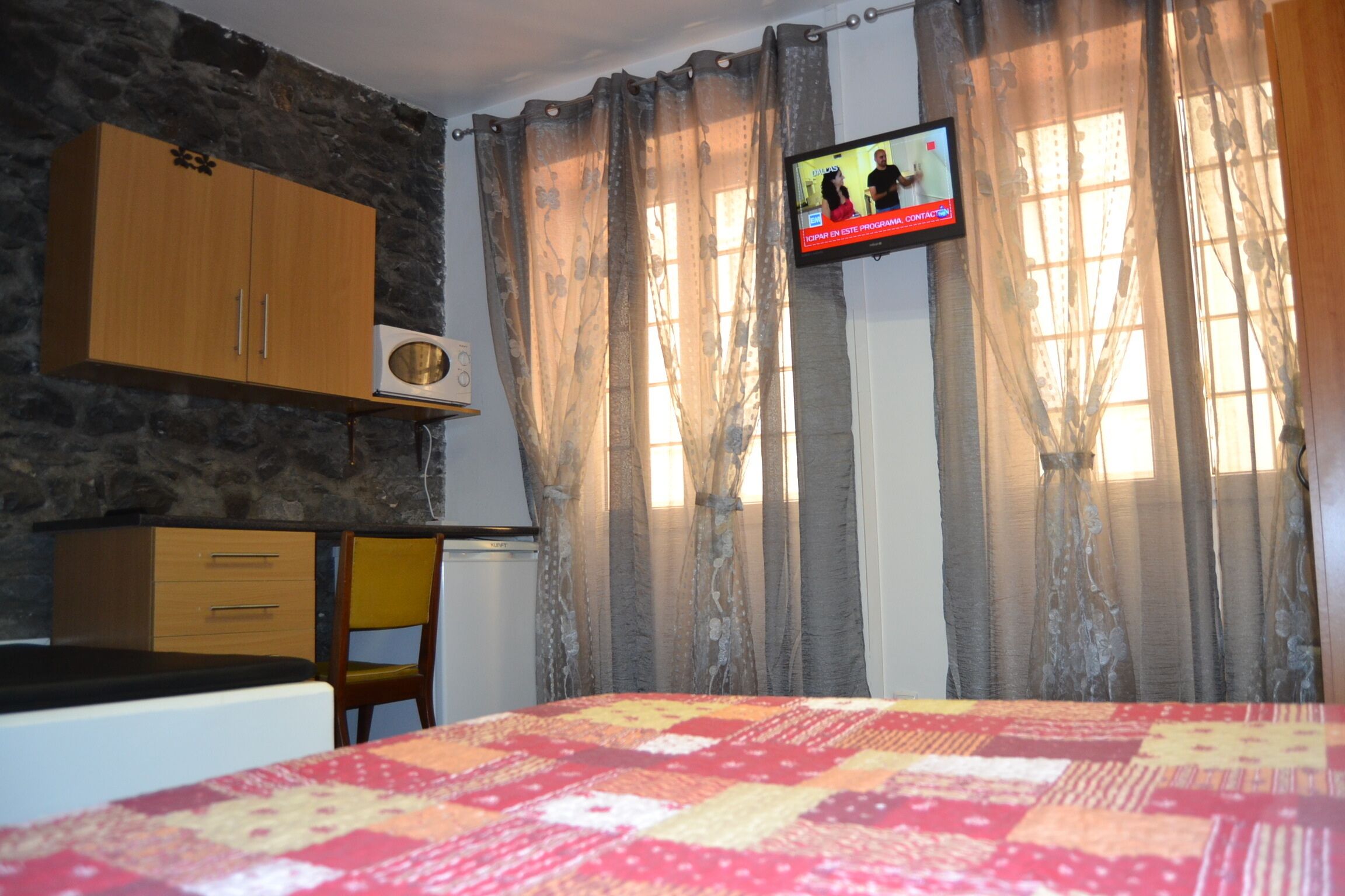A bedroom with a bed, a wooden wardrobe, a small table, a chair, and a television on the wall.