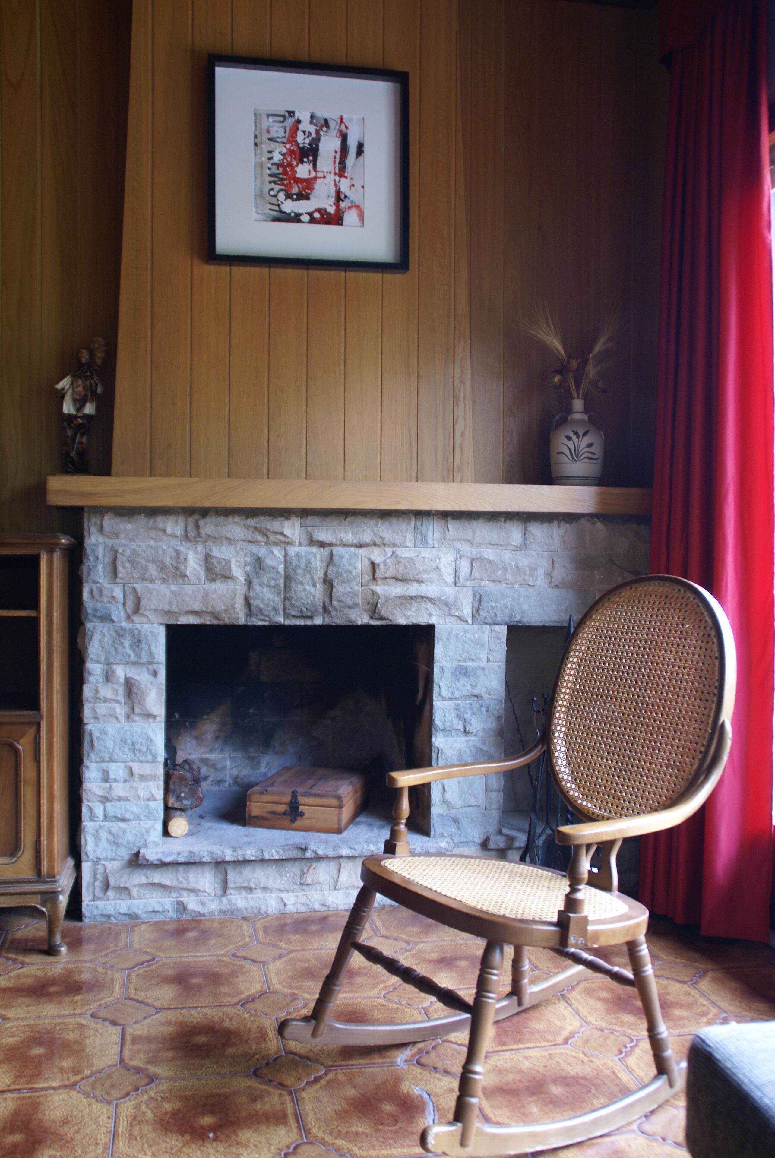 Un cuarto acogedor con una chimenea de piedra, un sillón mecededor y un cuadro enmarcado sobre la chimenea.