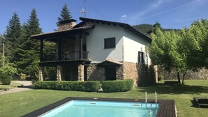 A two-story house with a stone foundation and a balcony, surrounded by a well-maintained lawn and a swimming pool.
