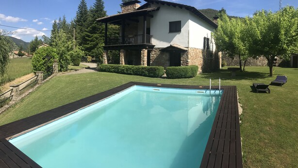 A swimming pool with a wooden deck, surrounded by a well-maintained lawn and a house with a balcony.