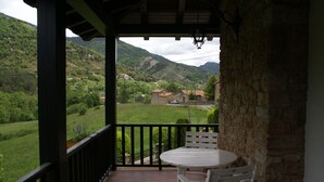 A balcony with a table and chairs, overlooking a green valley with houses and mountains.
