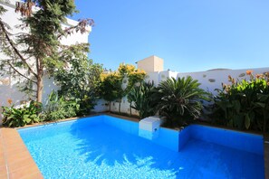 A rectangular swimming pool with a tiled edge, surrounded by various plants and a white building in the background. - Villa Kamilari (Faistos)