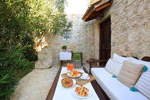 A stone-walled outdoor seating area with a wooden bench and a white sofa. A tray with food and drinks is placed on the table. - Villa Kamilari (Faistos)