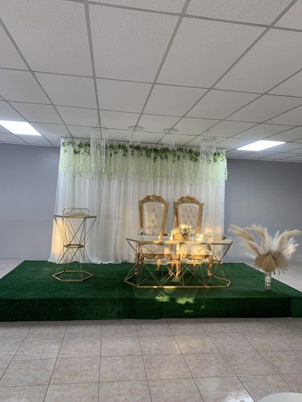 A stage with a green carpet, two chairs, a table, and a decorative backdrop. - Mi Tierra (Cabo Rojo)