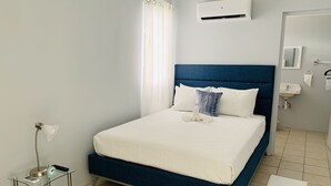 A neatly arranged hotel room with a bed, a bedside table with a lamp, a clock, and a small table with a remote control. - Mi Tierra (Cabo Rojo)