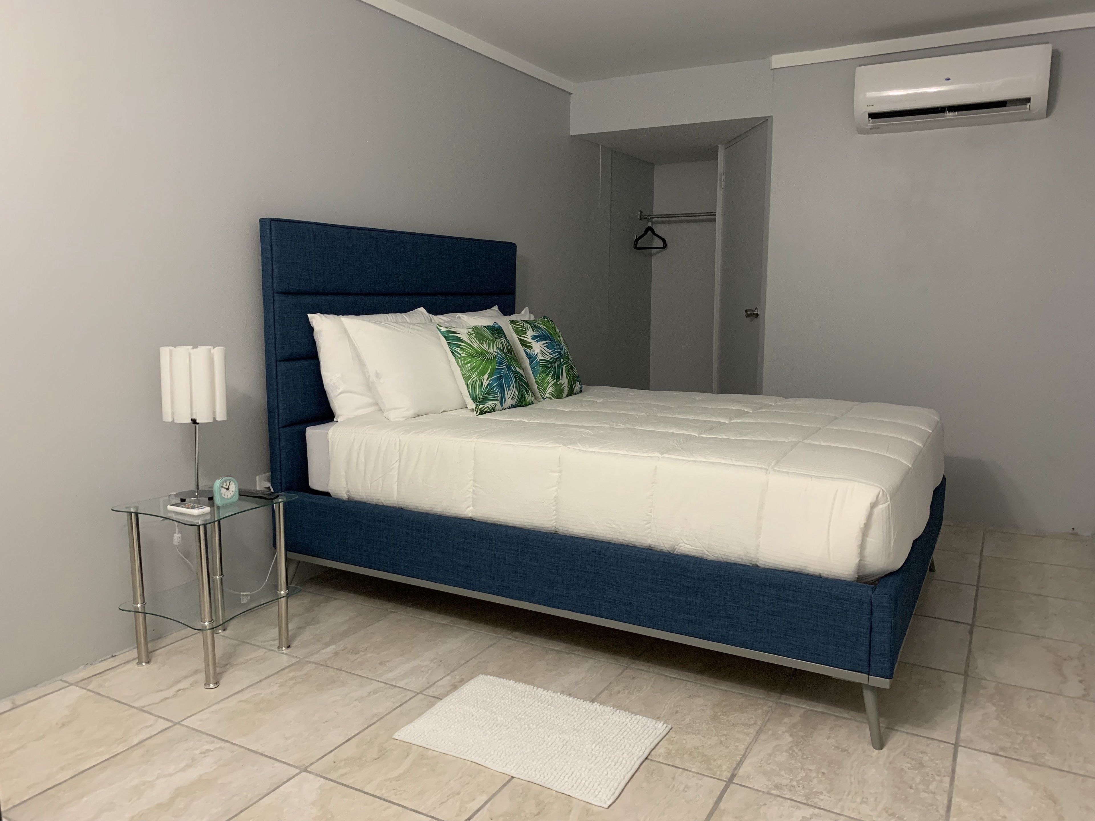 A modern bedroom with a blue upholstered bed, white bedding, a bedside table with a lamp, and a white rug.