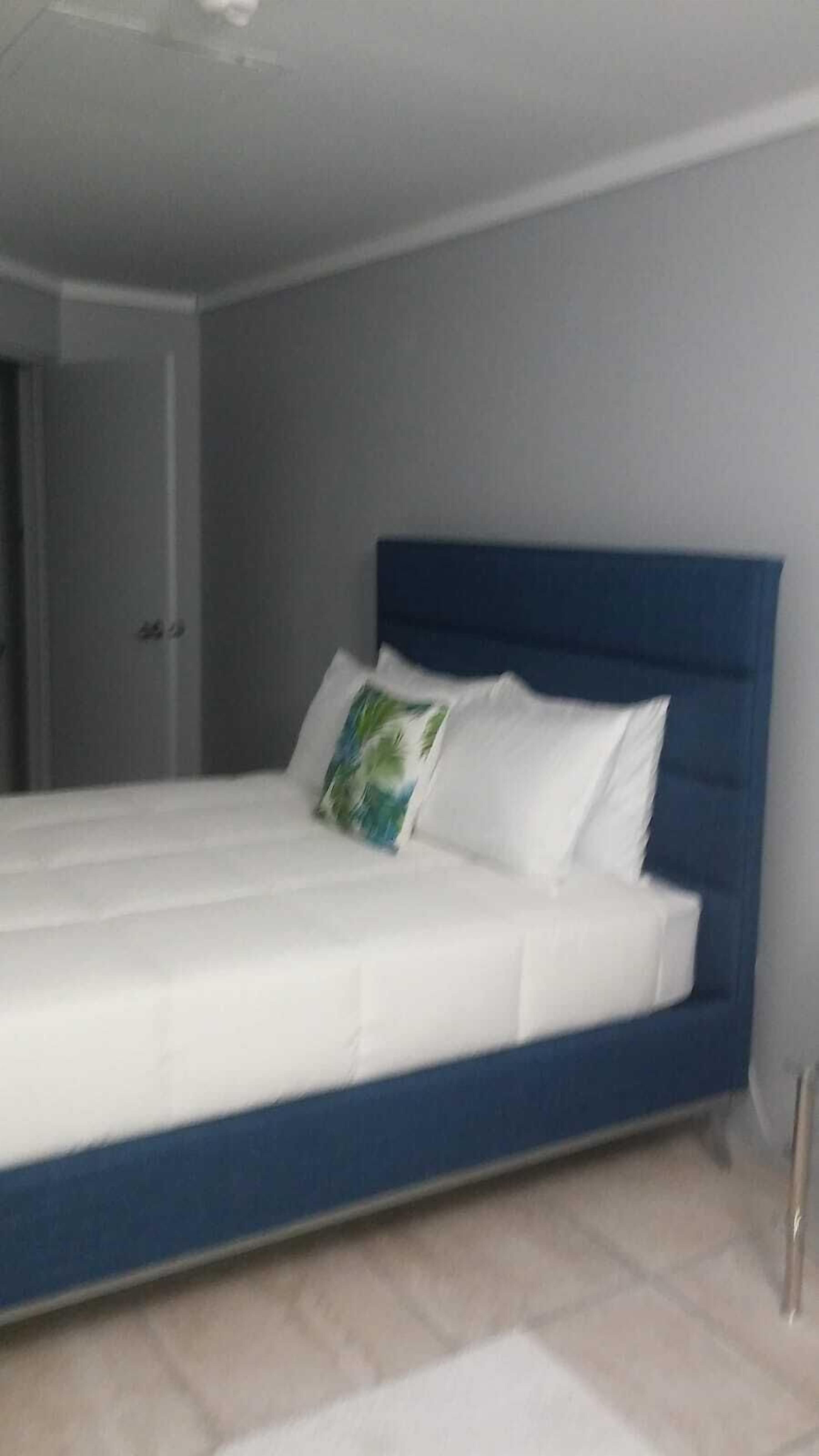 Una cama con cabecera azul y ropa de cama blanca en una habitación con paredes de tonos claros y suelo de baldosas.