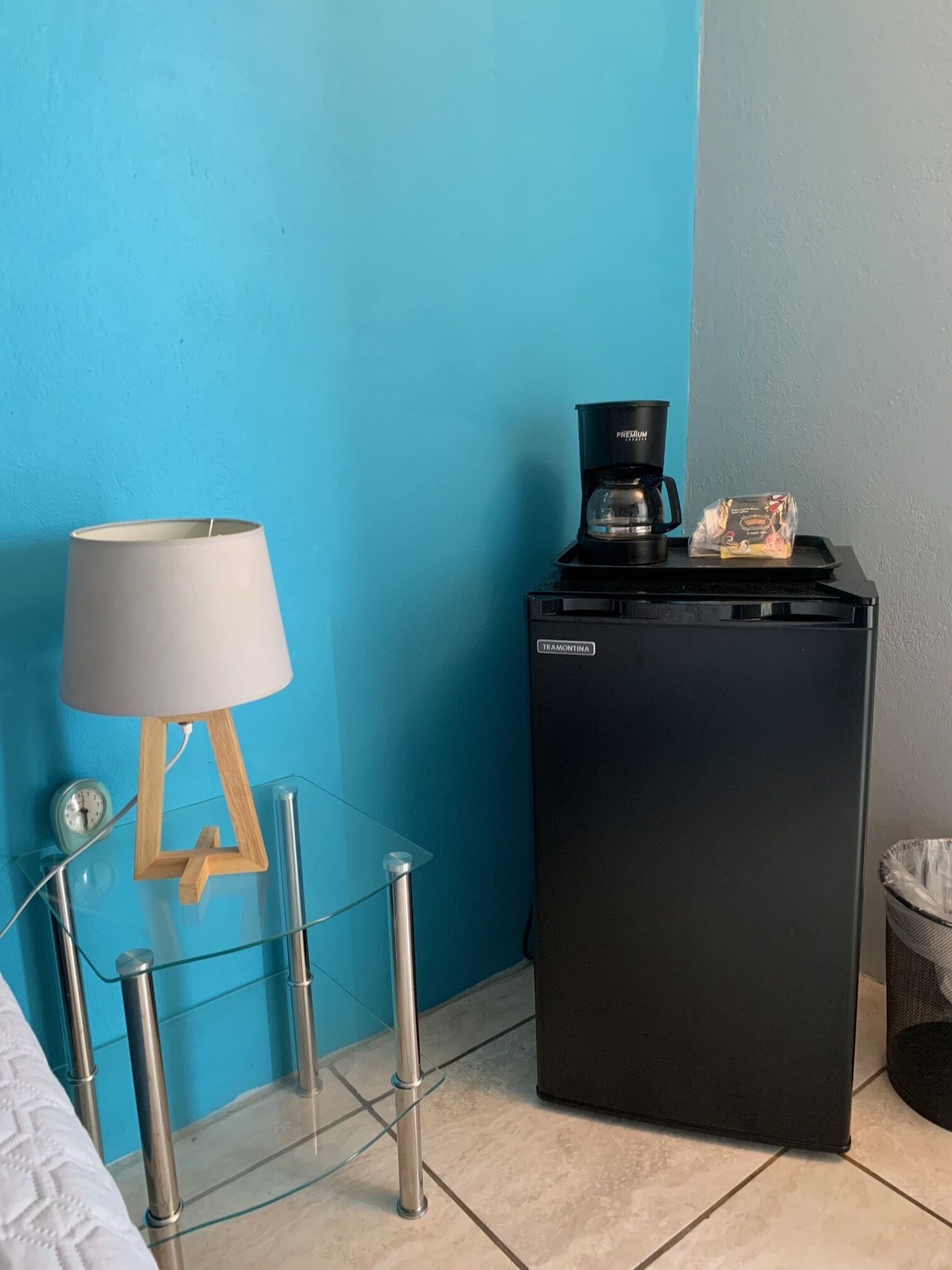 A compact room with a blue wall, a small black refrigerator, a coffee maker, a lamp on a glass table, and a trash bin.