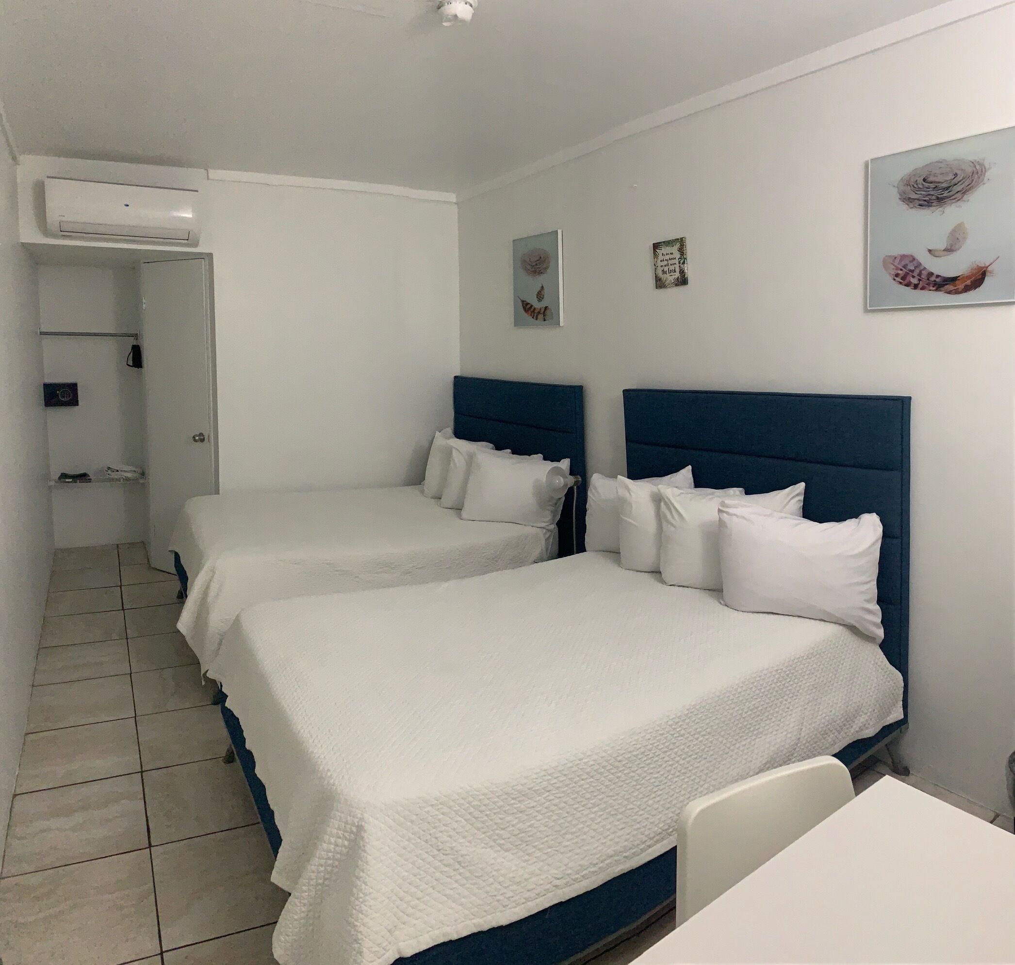 A hotel room with two beds, a white air conditioning unit, a small table, and artwork on the wall.