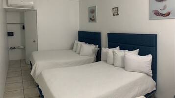 A hotel room with two beds, a white air conditioning unit, a small table, and artwork on the wall.
