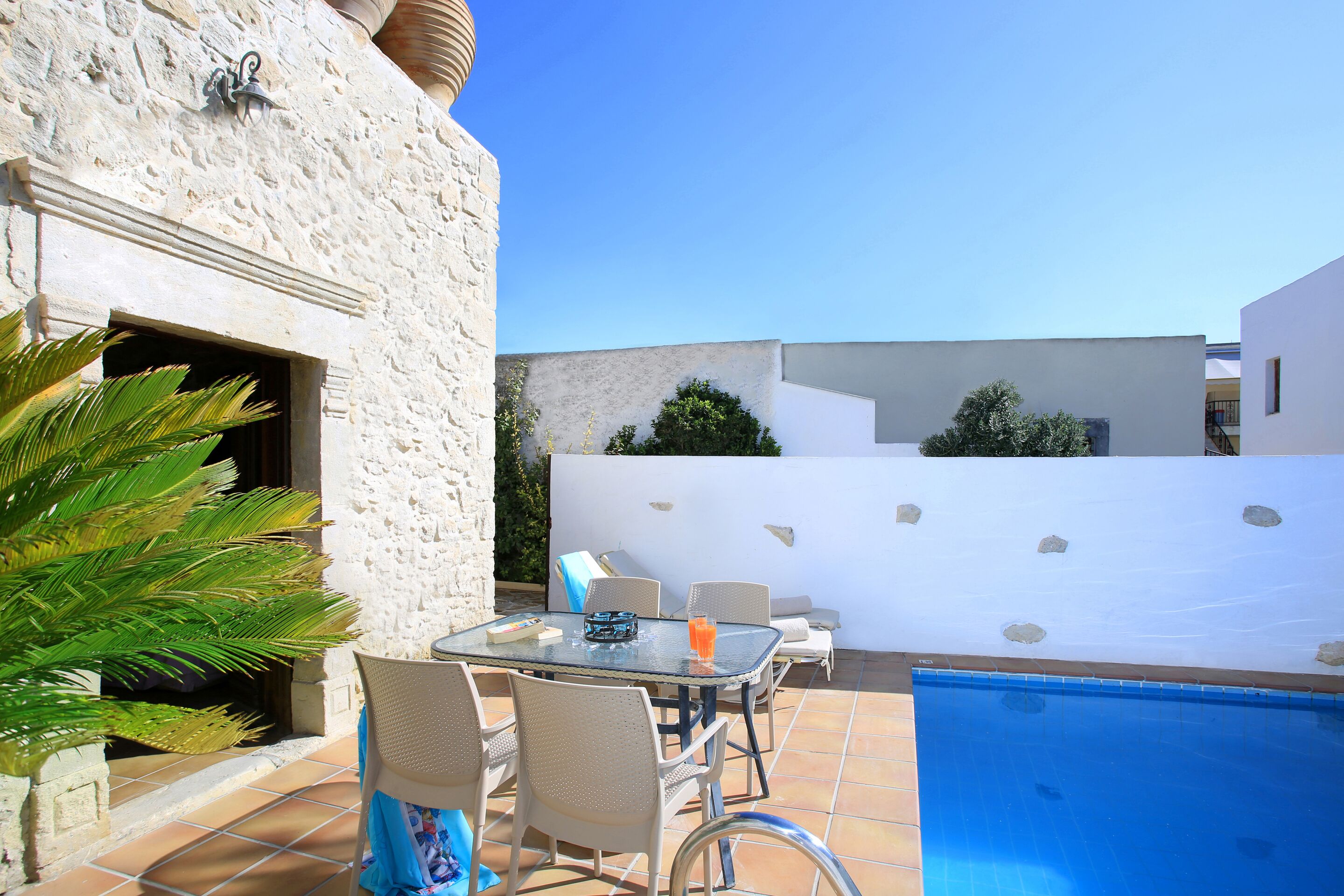 Un patio con una mesa y sillas de vidrio, una piscina y un muro de piedra.