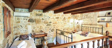 A rustic room with stone walls, wooden beams, a bed with a wooden headboard, a small table with a lamp, and a window with a view.