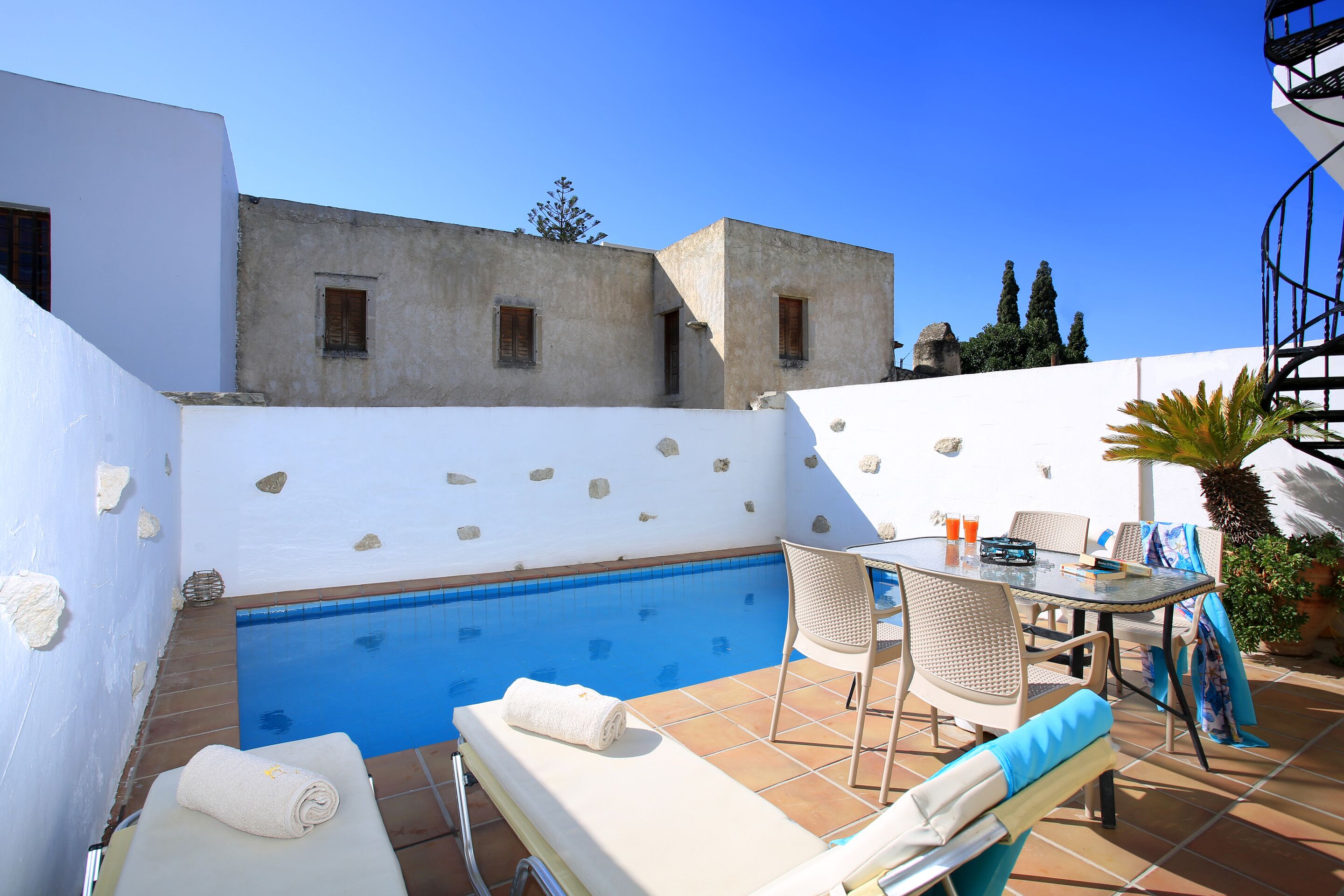 A poolside area with a white wall, a small pool, lounge chairs, and a table with chairs.