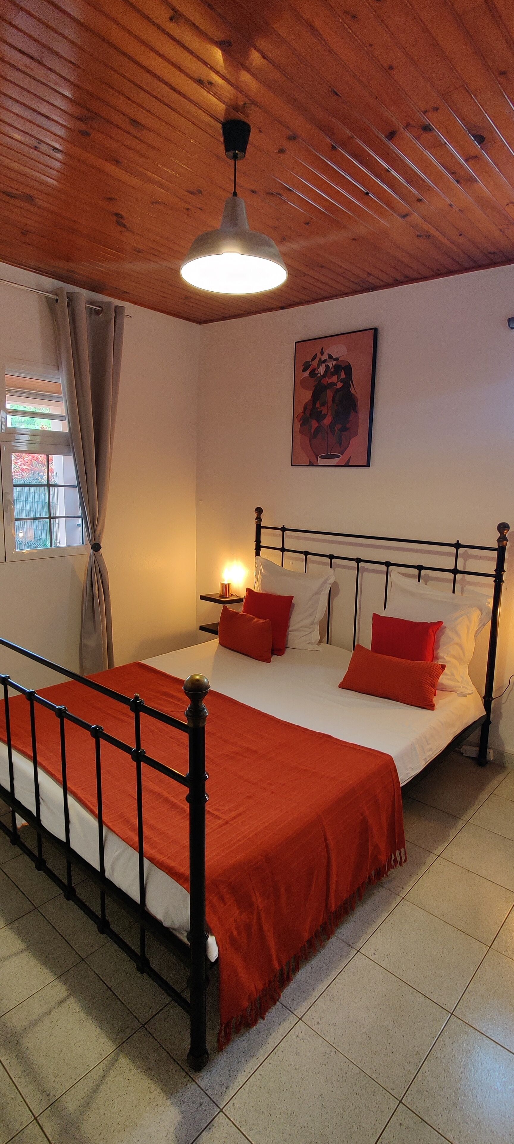 A bedroom with a wooden ceiling, a bed with red and white bedding, a bedside table with a lamp, and a window with curtains.
