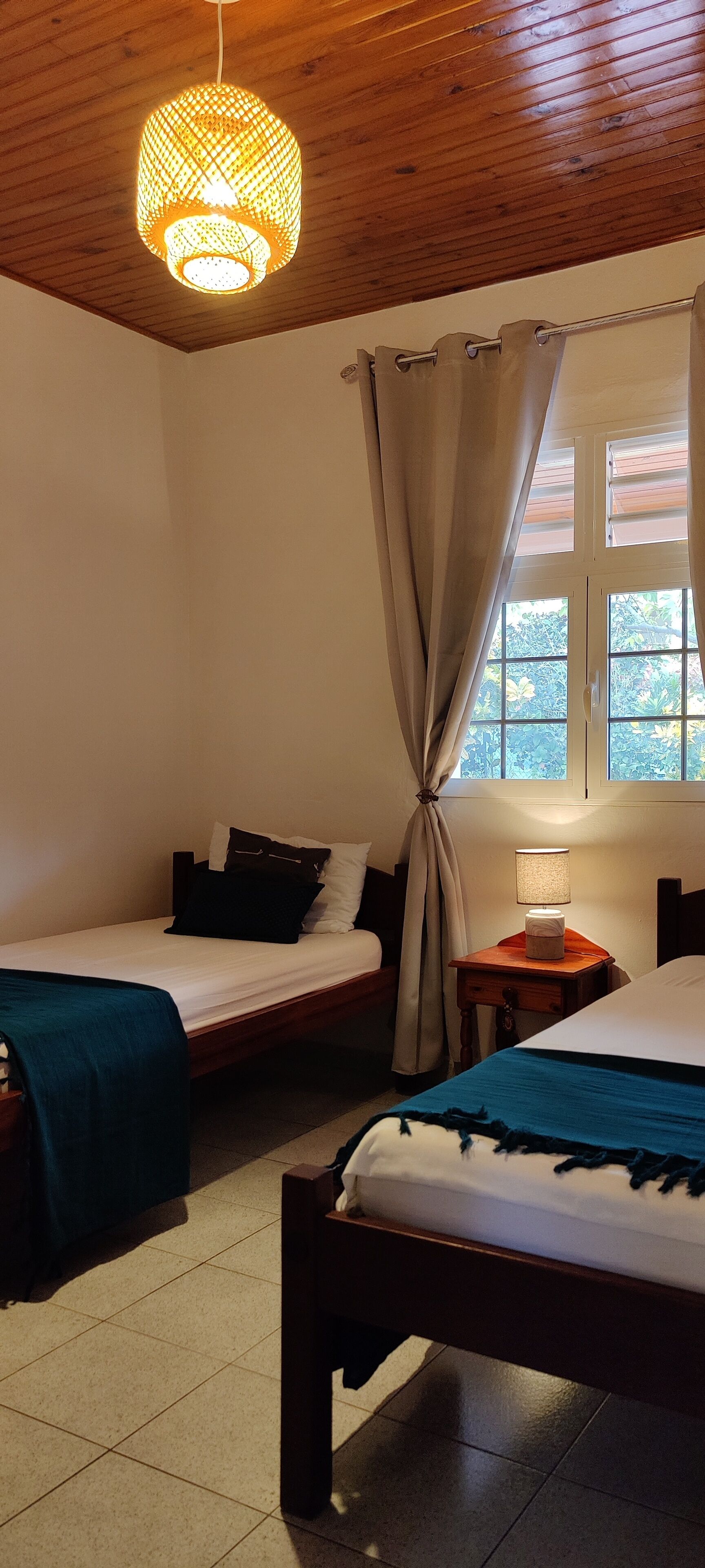 A room with two beds, a wooden ceiling, a window with curtains, and a nightstand with a lamp.