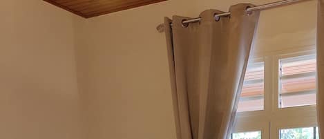 A room with two beds, a wooden ceiling, a window with curtains, and a nightstand with a lamp.