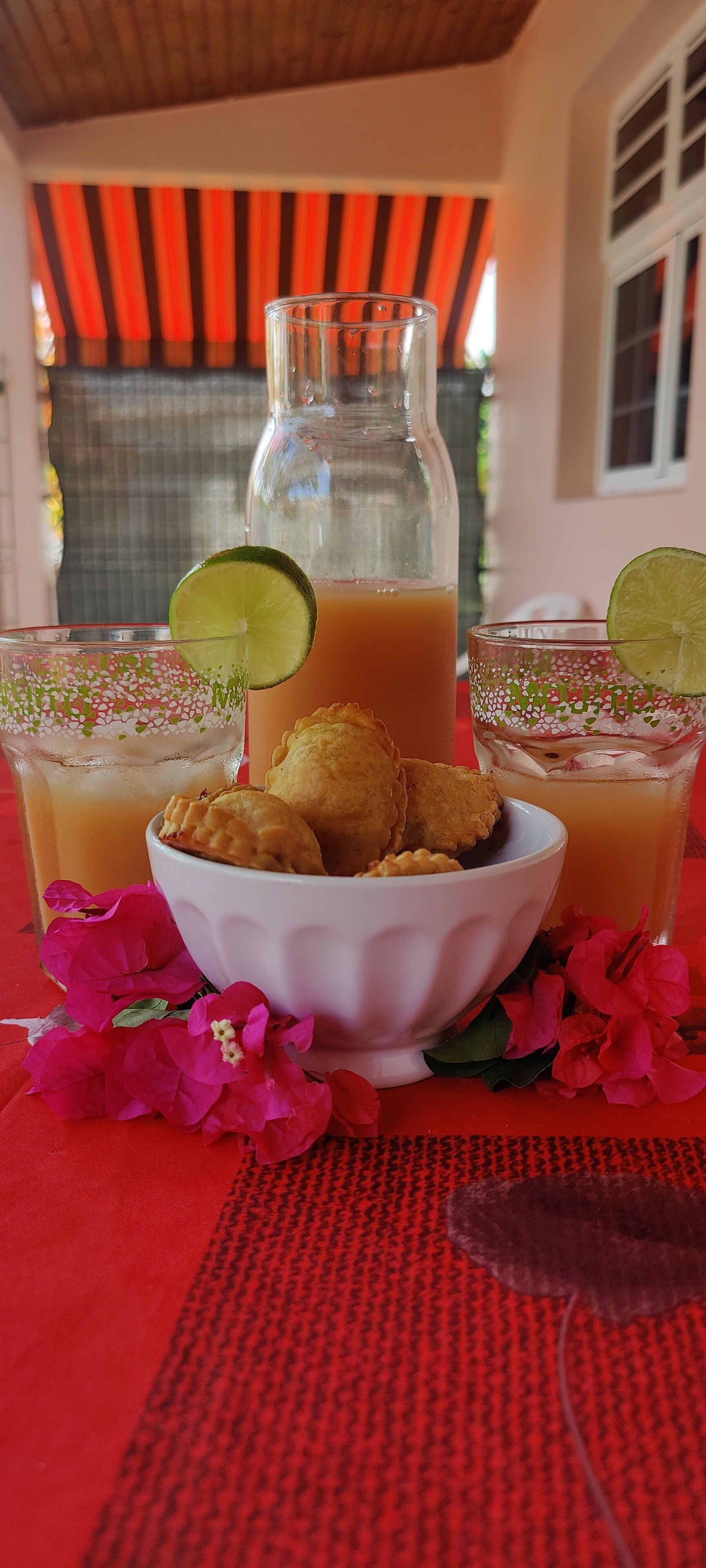 Une table avec un pichet de jus d’orange, deux verres de jus d’orange, un bol de grignotages frits et des fleurs roses.