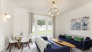 Een moderne woonkamer met een blauwe bank, witte stoelen, een houten salontafel en een eettafel met een fles en glazen.