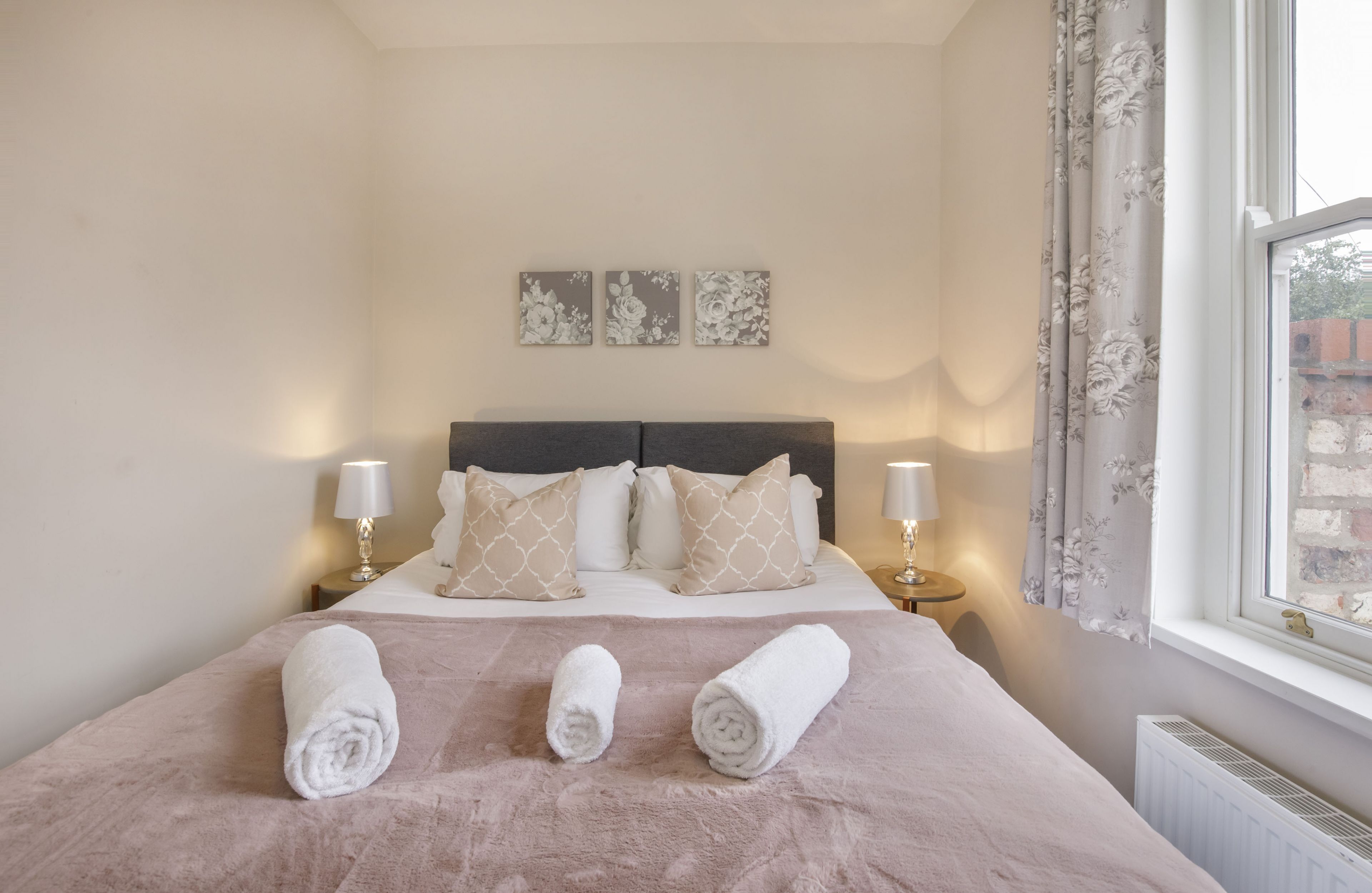 Ein ordentlich bezogenes Bett mit einer rosa Decke und drei zusammengerollten Handtüchern, zwei Nachttische mit Lampen sowie ein Fenster mit Vorhängen.