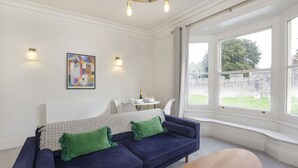 Een moderne woonkamer met een blauwe bank, groene kussens, een houten salontafel en een groot raam met uitzicht op de omgeving.