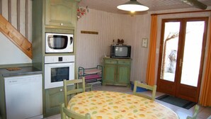 Uma cozinha compacta com micro-ondas, forno e uma pequena televisão sobre um armário. Há uma mesa de jantar com cadeiras e uma porta que dá para o exterior.