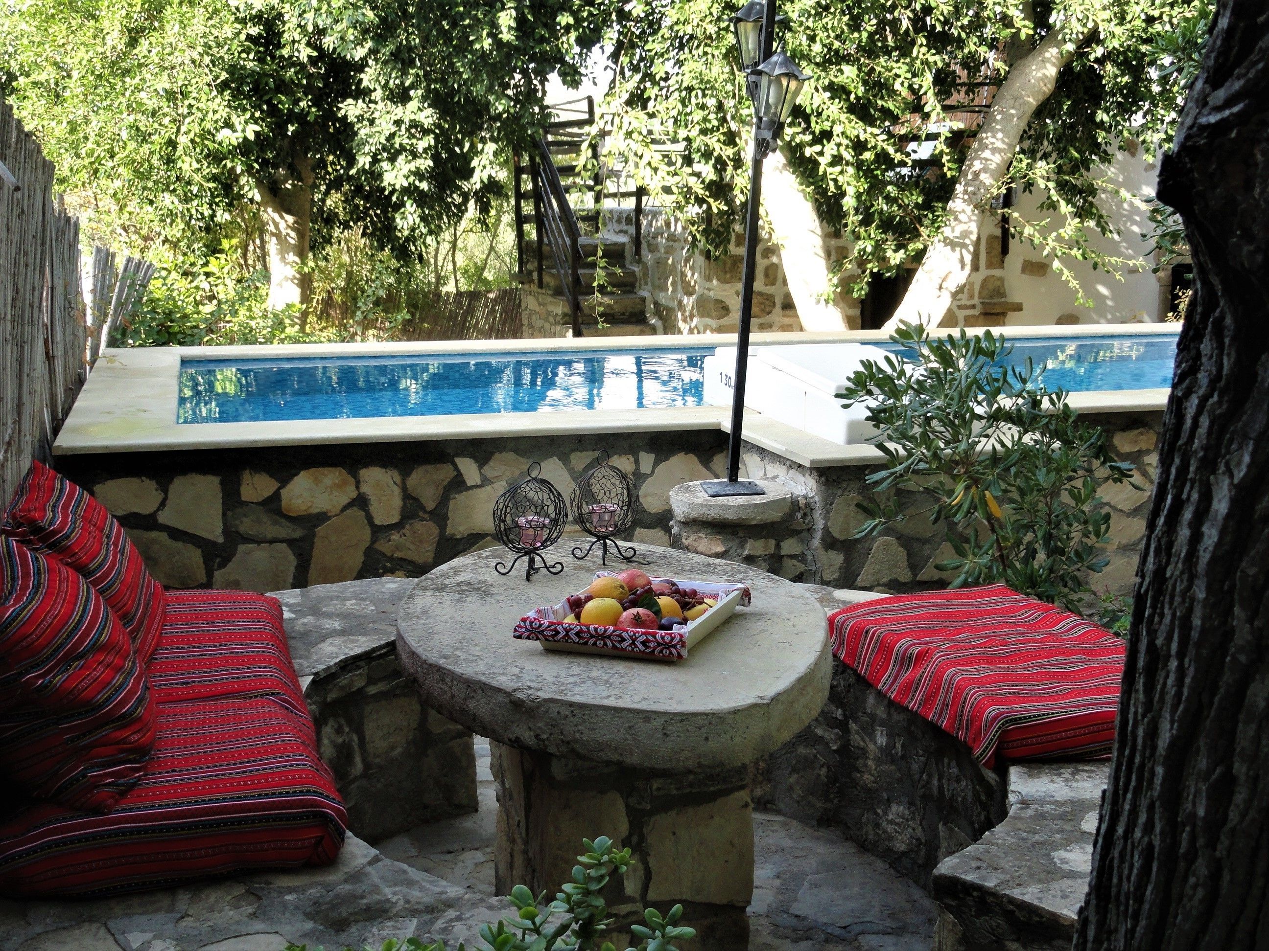 A stone patio with a round table, red patterned cushions, and a fruit basket. A stone wall and a pool are visible in the background.