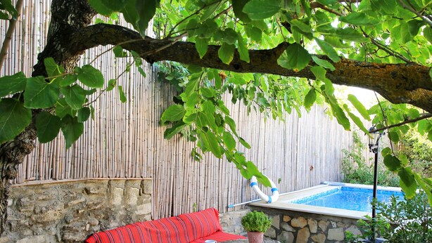 Eine Steinterrasse mit einem rotgestreiften Sofa und Kissen, einem kleinen Tisch mit einem Blumentopf und Blick auf einen Swimmingpool im Hintergrund.